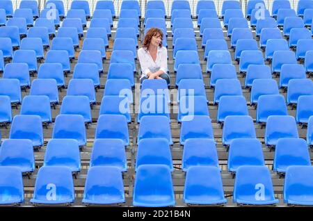 Donna glum seduta in file vuote di posti a sedere spettatore blu in un auditorium o in uno stadio con un'espressione annoiata Foto Stock