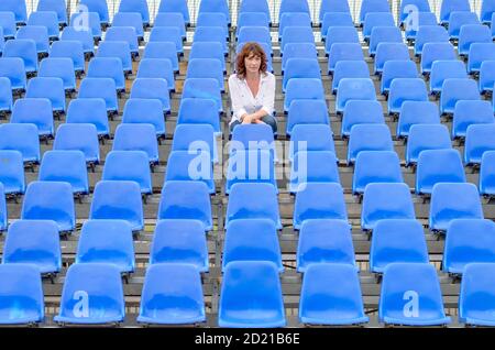 Donna glum seduta in file vuote di posti a sedere spettatore blu in un auditorium o in uno stadio con un'espressione annoiata Foto Stock
