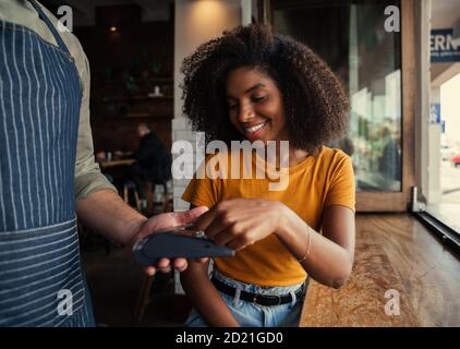 Sorridente donna da corsa mista con afro che paga felicemente il caffè alla caffetteria. Foto Stock