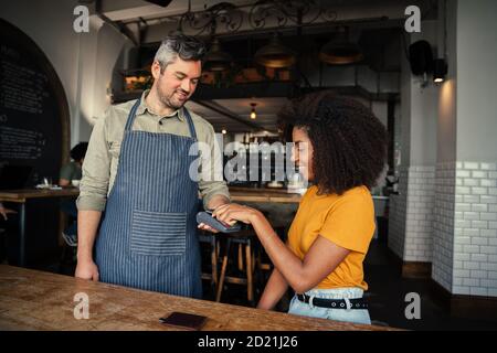 Bella donna etnica che tap carta facendo il pagamento per il caffè al bel cameriere in un bar alla moda. Foto Stock