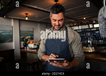 Il proprietario di una caffetteria maschile sorride al tablet mentre si trova nella caffetteria. Foto Stock