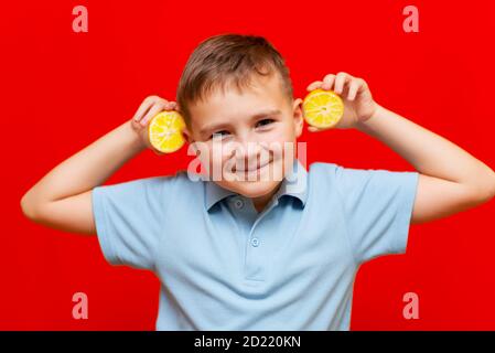 Persone sane. Divertente immagine sorridente del ragazzo della scuola che mostra due limoni vicino alla testa . Isolato su sfondo rosso alla moda Foto Stock