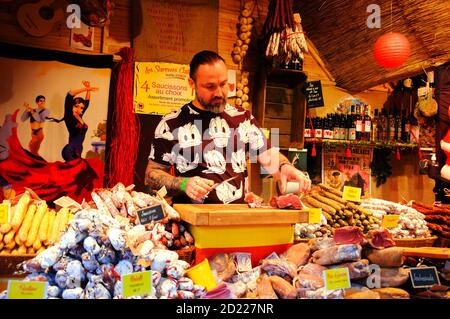 PARIGI, FRANCIA - 26 NOVEMBRE 2017: Macellaio giovane in divertente t-shirt che vende prosciutto tradizionale catalano e salsicce secche al mercato di Natale a la Defense Foto Stock