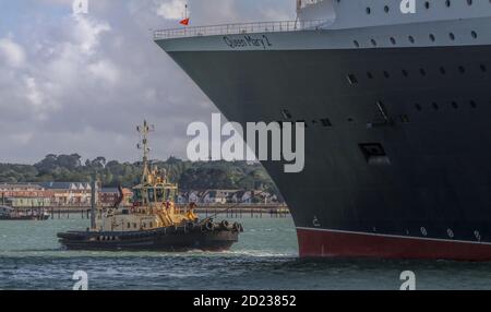 Potente piccolo Tugboat che guida, tirando e trainando la nave da crociera Queen Mary Two via da UN ormeggio mentre si prepara a partire. Southampton Docks Regno Unito Foto Stock