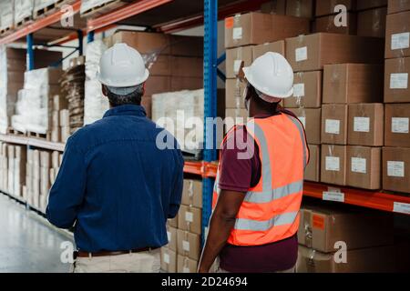 Uomo d'affari caucasico e lavoratore di fabbrica etnico che cammina attraverso il processo di imballaggio per le consegne in magazzino. Foto Stock