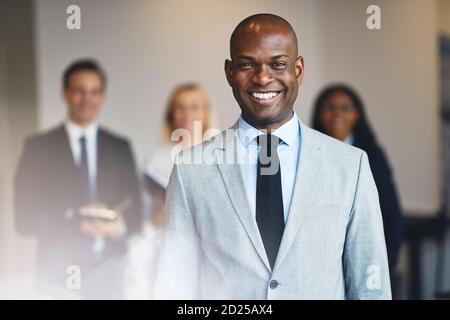 Sorridente giovane uomo d'affari africano in piedi in un ufficio con diversi colleghi in piedi sullo sfondo Foto Stock