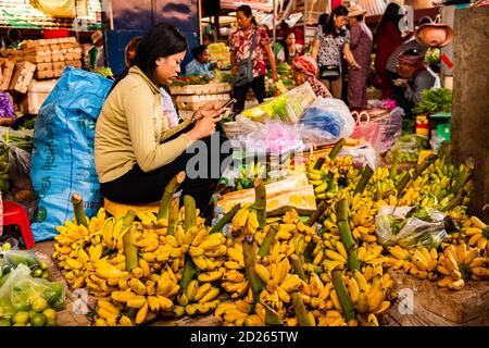 KAMPOT, CAMBOGIA - Agosto 14, 2018: Kampot, Cambogia, Agosto 2019 - UNA donna commerciante che usa il suo telefono mentre vende verdure e frutta al Kampot fresco ma Foto Stock