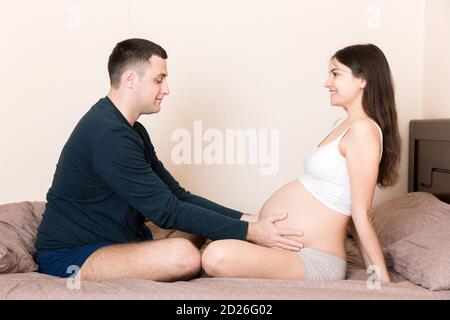 Donna incinta con marito seduto a letto, tenendo le mani sul ventre a casa. Foto Stock