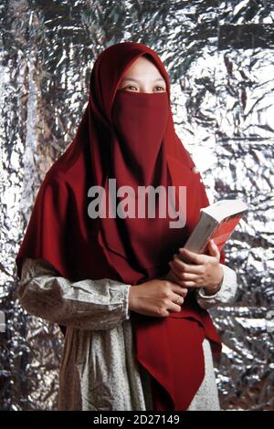 Un ritratto di una donna asiatica velata che tiene un libro. Bel modello musulmano che indossa abiti rossi. Closeup di uno studente islamico Foto Stock