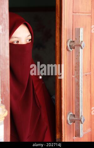 Ritratto di una donna asiatica velata che apre la porta di una casa. Bel modello musulmano che indossa abiti rossi Foto Stock