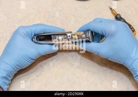 Le mani maschili in guanti blu riparano una sigaretta elettronica su un tavolo luminoso in legno da vicino Foto Stock