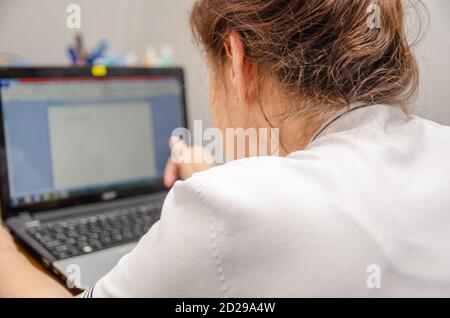 La donna anziana dai capelli scuri è seduta a una scrivania digitando su un computer portatile, foto vista posteriore Foto Stock