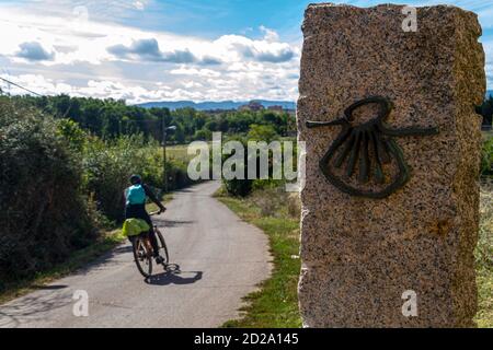 Ciclista pellegrina sulla rotta del Camino de Santiago (Via di San Giacomo) passando accanto allo shell dello scaloppo che segna la via Foto Stock