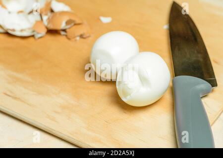 Su una tavola di legno ci sono due uova di pollo sbucciate per affettare e un coltello Foto Stock