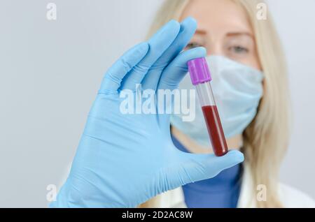 giovane donna bionda medico tiene una provetta con sangue in mano in guanti blu da vicino. Concetto di campionamento del sangue, ricerca medica e chimica Foto Stock