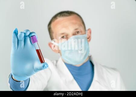 Il medico di sesso maschile in un abito medico, maschera e guanti contiene una provetta con sangue. Concetto di test medici Foto Stock