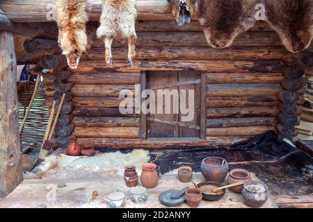 Vecchia casa in legno in stile rurale con pelli appese. Ingresso alla capanna, pelli animali sul pavimento e ceramiche d'epoca. Foto Stock