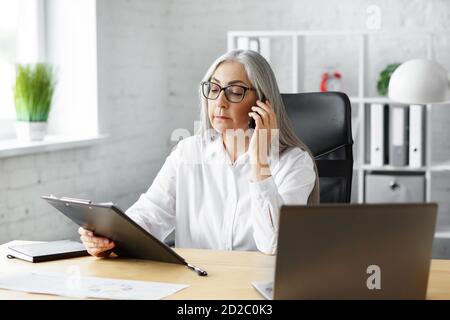 Donna anziana dai capelli grigi bella con smartphone e sorridente. Donna d'affari felice che usa le applicazioni del telefono cellulare, SMS, navigare in internet Foto Stock