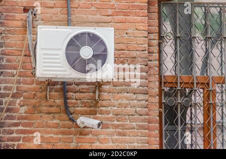 Aria condizionata bianca sulla parete di mattoni dell'edificio vicino alla finestra con bar, sotto l'aria condizionata è una telecamera di sorveglianza Foto Stock