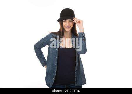 Donna cute ragazza che tocca il suo cappello nero in segno di saluto, salutando qualcuno che dice hi con il gesto della mano isolato su bianco sfondo della parete dello studio Foto Stock