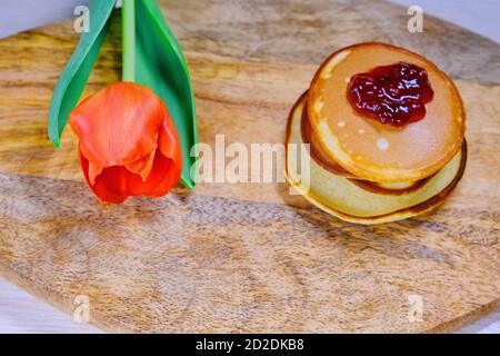 Pancake con marmellata e tulipano. Un fiore rosso si trova accanto ai dolci sulla tavola della cucina. Una collina di frittelle fresche sul tavolo in primavera. Foto Stock