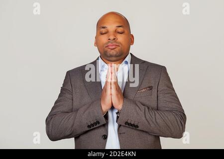 Bel giovane uomo d'affari afro-americano concentrato in giacca tenendo le mani insieme di fronte al viso, gli occhi chiusi pregando e sperando oggi è un buon giorno, su sfondo grigio Foto Stock