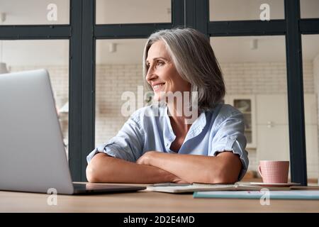 Sorridente donna matura di mezza età sognando guardando via al posto di lavoro. Foto Stock