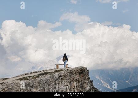 Una donna con uno zaino sul bordo di una roccia nelle Dolomiti, in Italia, vicino ad una panchina Foto Stock