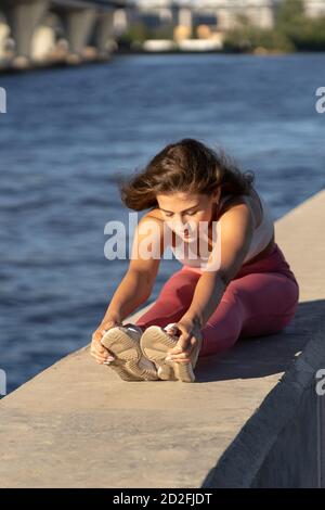 Giovane corridore yoga in gamba rosa seduta sull'argine, che allunga i muscoli in posizione seduta in avanti curva. Esercizi di riscaldamento stretch di Hamstring Foto Stock