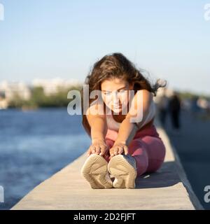 Donna giovane in forma con gamba rosa seduta sull'argine, muscoli tendenti in posizione seduta in avanti curva, fuoco selettivo. Riscaldamento stretch Hamstring Foto Stock