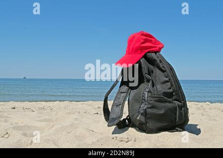 Zaino nero con due cinghie e su di esso un cappuccio rosso sulla riva della sabbia grigia sciolta dai piedi del mare blu liscio e calmo. Foto Stock