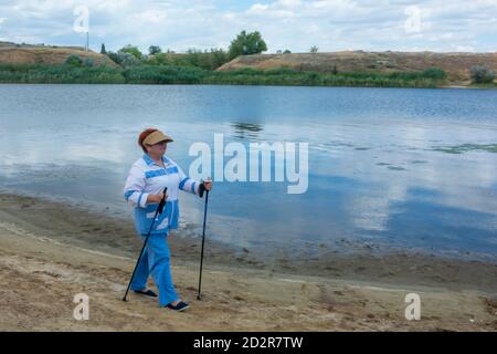 Scandinavo a piedi, una donna anziana cammina lungo la riva del fiume con bastoni. Concetto di stile di vita attivo nella vecchiaia. Messa a fuoco morbida, sfocatura. Foto Stock