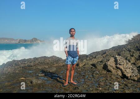 Onde che colpiscono rocce rotonde e spruzzi. Un giovane si erge su una riva rocciosa e le onde si infrangono contro una scogliera Foto Stock