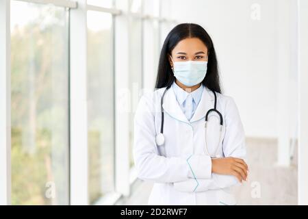 Medico femminile che indossa una maschera medica con le braccia incrociate in background ospedaliero. Medicina, professione e concetto sanitario Foto Stock