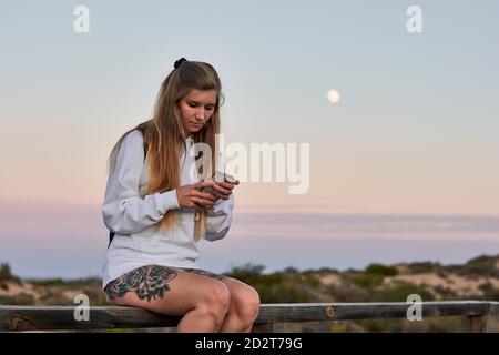 Tranquilla viaggiatore femminile seduta su recinzione di legno sul lungomare e. messaggi sui social media tramite cellulare mentre ci si rilassa al tramonto in estate Foto Stock