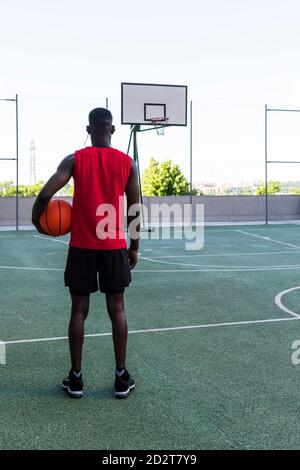 Vista posteriore del giocatore africano americano maschio in piedi sportswear con pallacanestro nel parco giochi e vista lontano Foto Stock