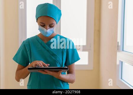 Giovane infermiera in scrub blu e maschera medica che legge le informazioni su tablet mentre si trova nel corridoio dell'ospedale moderno Foto Stock