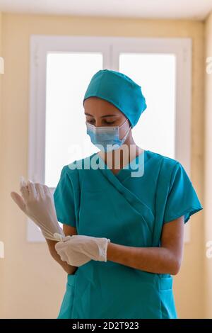 Giovane infermiera in scrub blu e maschera medica che si mette guanti sterili in lattice durante la preparazione per il lavoro in clinica durante pandemia di coronavirus Foto Stock