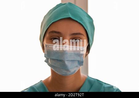 Donna medico indossando maschera medica e uniforme in piedi in clinica Vicino alla finestra e guardando la fotocamera durante la pandemia COVID 19 Foto Stock