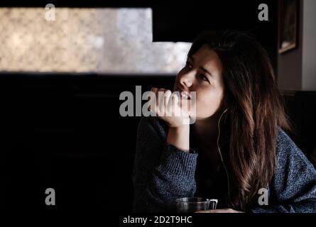 Sognante donna seduta al tavolo nel caffè e ascoltata musica nelle cuffie mentre si appoggia a mano e si guarda con attenzione via Foto Stock