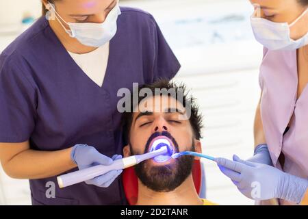 Dentista femminile e assistente che usa la luce e l'aspirazione di cura dentale dispositivo sui denti del maschio con gli occhi chiusi clinica Foto Stock