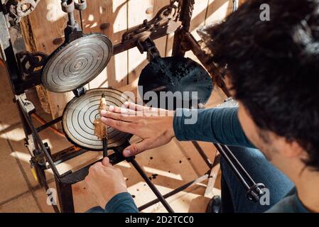 Dall'alto, un artigiano irriconoscibile tagliato utilizzando il bastone durante l'arrotolamento Waffle catalani caldi appena sfornati al neula sul ferro da stiro a base di waffle fiamma sopra gas Foto Stock