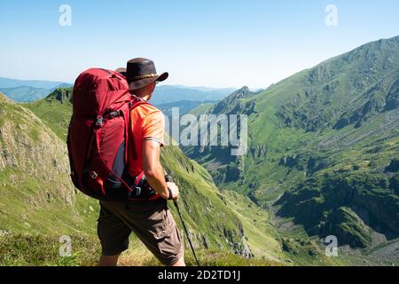 Vista posteriore di escursionista maschile irriconoscibile con zaino e trekking rimanere in piedi sulla collina erbosa e ammirare lo spettacolare scenario di cresta di montagna in estate giorno Foto Stock