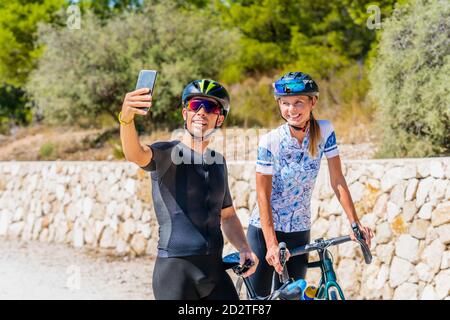 Felice giovane uomo e donna attivi in abbigliamento sportivo e caschi protettivi prendere selfie su smartphone mentre si è in piedi con le biciclette sulla strada di campagna e prepararsi per la guida Foto Stock