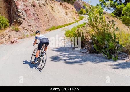 Vista posteriore a lunghezza intera di un'anonima forte corsa in bicicletta femminile pedala su una strada asfaltata curvilinea che salirà tra le montagne Foto Stock