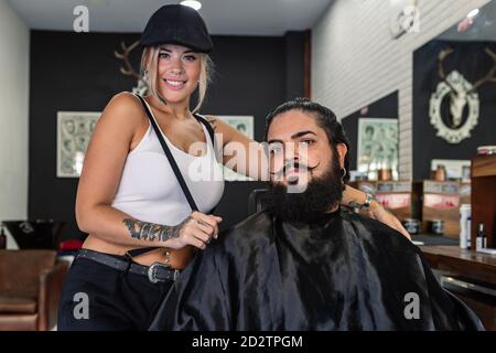 Allegro hipster tatooed femmina in piedi vicino al cliente di sesso maschile bearded con rifinitura elegante dei capelli e dei baffi e guardare la fotocamera mentre si lavora in barbiere professionale Foto Stock