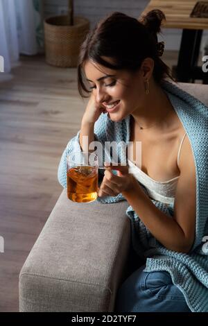 Giovane donna pusiva avvolta in coperta calda seduta sul divano e gustando il tè caldo mentre si rilassa nell'accogliente soggiorno a casa Foto Stock