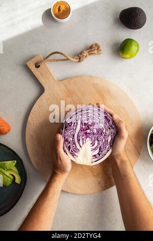 Vista dall'alto della persona anonima del raccolto che mette a metà il rosso fresco cavolo su tagliere di legno mentre preparano l'insalata deliziosa dentro cucina Foto Stock