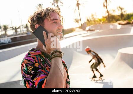 Vista laterale di un ragazzo con capelli ricci millenni in stile outfit che parla sul telefono cellulare mentre ci si trova vicino a una rampa di cemento in skatepark all'aperto durante il giorno d'estate Foto Stock
