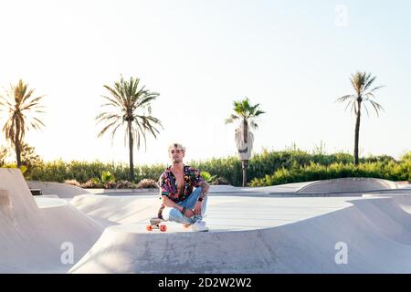 Basso angolo di giovane elegante skater maschile in camicia colorata e jeans seduti a skateboard mentre si passa la giornata estiva in skatepark Foto Stock